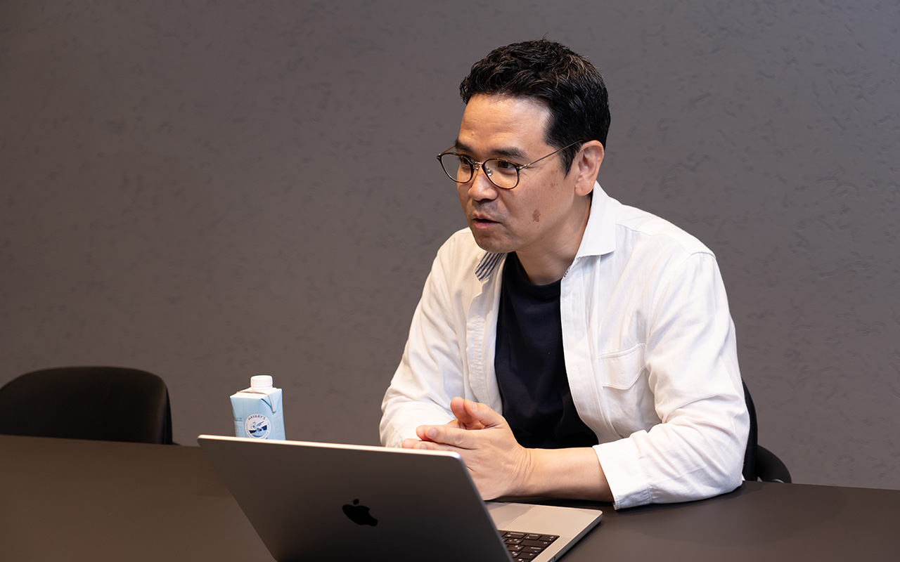 田中が机に置かれたノートパソコンに向かい、両手を軽く組みながら前傾姿勢で話している様子