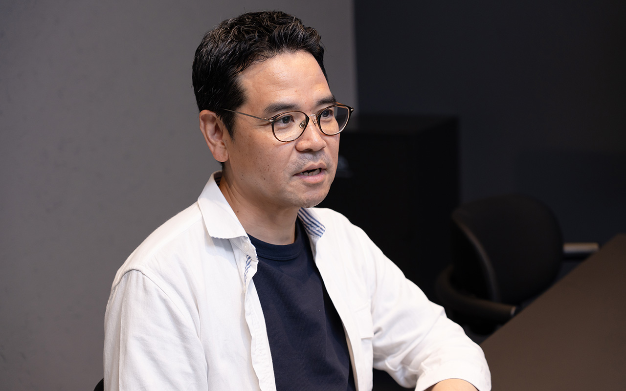 田中がやや横を向きながら口を開いて話しており、真剣な表情で相手に視線を向けている様子