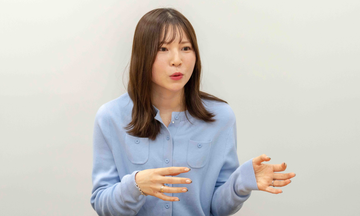 功刀の写真。淡い水色のカーディガンを着ており、肩あたりまでのまっすぐな髪が自然に流れている。口を少し開き、話している最中のような表情で、両手を前に出して指を軽く開きながら説明する仕草をしている。