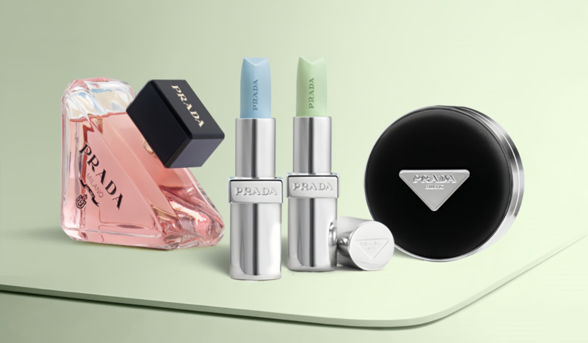 淡いグリーンの背景の上に、PRADAのコスメとフレグランスが並んでいる。左にはピンク色の液体が入った香水ボトルが斜めに置かれ、黒いキャップが付いている。中央にはシルバーのケースに入った2本のリップが立てて並び、片方は薄いブルー、もう片方は淡いグリーンの色味をしている。手前にはリップのキャップが横向きに置かれている。右側には黒いラウンド型のコンパクトが配置され、中央にPRADAのロゴが入っている。