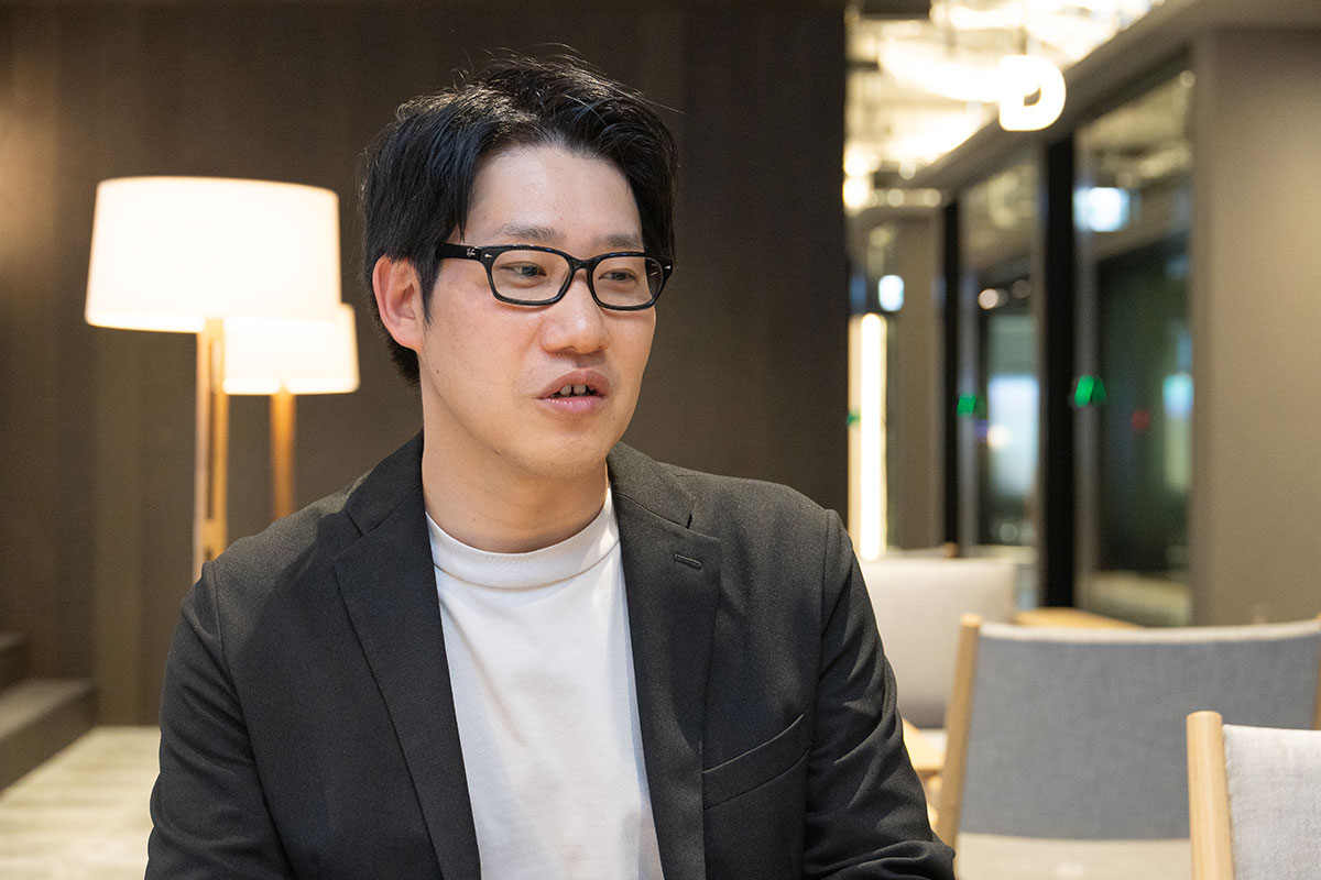 林が椅子に座り、横を向いて話している写真。黒縁眼鏡と黒いジャケットを着用し、落ち着いた表情で会話に集中している。背景には照明やラウンジ空間が広がる。