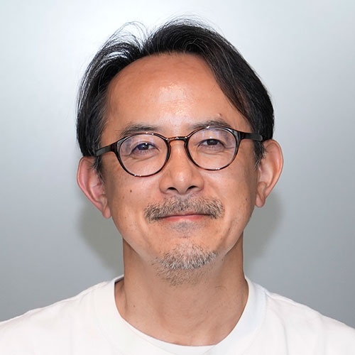 丸眼鏡をかけた川邊のバストショット。白いTシャツを着用し、口ひげとあごひげを整えて穏やかに微笑んでいる。グレーの背景で撮影されたポートレート写真。