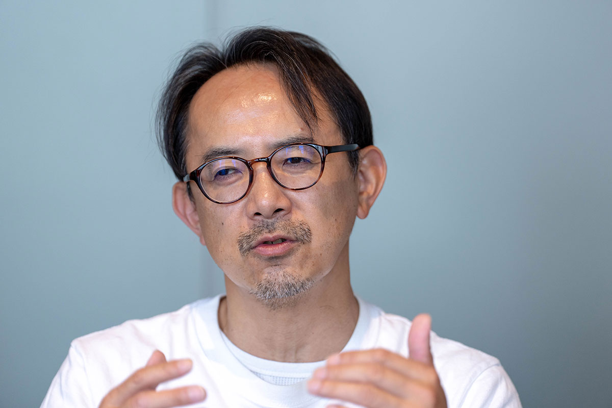 白いTシャツ姿の川邊が両手を上げながら話している写真。丸眼鏡をかけ、真剣な表情で説明している。背景は淡い青色で、上半身が写っている。