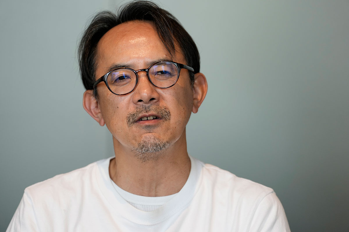 白いTシャツを着た川邊の上半身写真。丸眼鏡と口ひげが特徴で、正面を向きながら落ち着いた表情で話している。背景は淡い青色である。