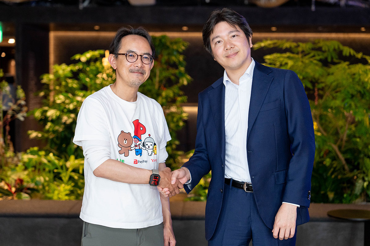 白いTシャツ姿の川邊とネイビーのスーツ姿の妹尾が握手を交わす写真。屋内の緑豊かな空間で並んで立ち、カメラに向かって微笑んでいる。
