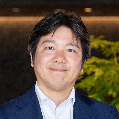 ネイビーのスーツを着た妹尾のバストショット。黒髪を整え、穏やかな表情でカメラを見つめている。背景には緑の植物がぼけて写る。