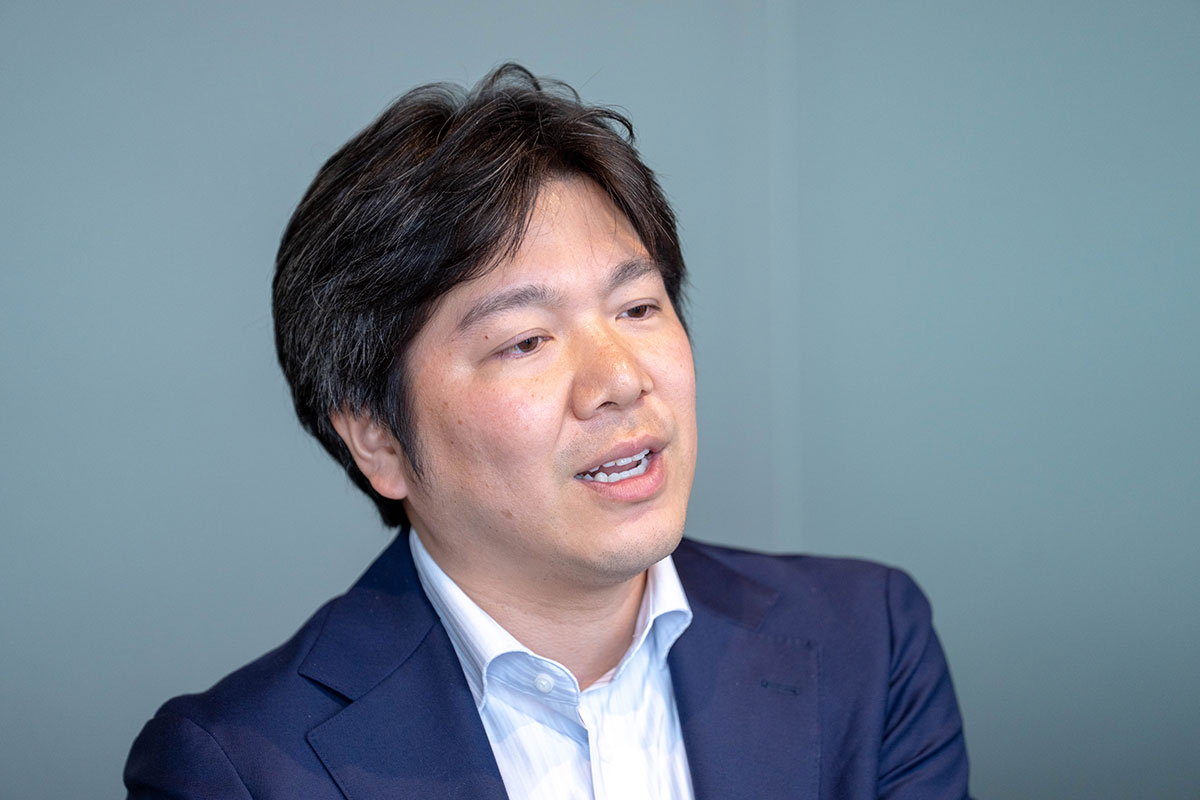 ネイビーのスーツ姿の妹尾が話している写真。横を向きながら口を開いて説明し、落ち着いた表情で会話している。背景は淡い青色である。