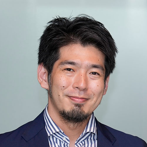 ストライプ柄のシャツにネイビーのジャケットを着た菅野先生のバストショット。口ひげとあごひげが特徴で、微笑みながらカメラを見ている。