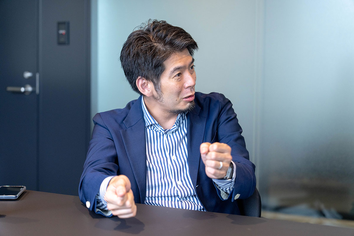 会議室で菅野先生が両手を前に出しながら話している写真。ストライプ柄のシャツとネイビーのジャケット姿で、身振りを交えて説明している。