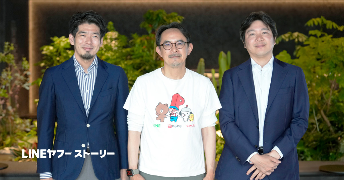 3人の男性が屋内の緑豊かな空間で並んで立つ写真。中央の川邊はキャラクターが描かれた白いTシャツを着用し、左右の男性はネイビーのスーツ姿でカメラに向かって微笑んでいる。左下に「LINEヤフーストーリー」の文字がある