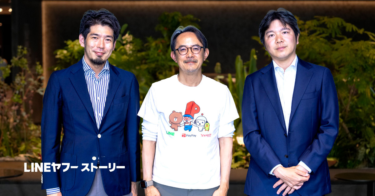 3人の男性が屋内の緑豊かな空間で並んで立つ写真。中央の川邊はキャラクターが描かれた白いTシャツを着用し、左右の男性はネイビーのスーツ姿でカメラに向かって微笑んでいる。左下に「LINEヤフーストーリー」の文字がある