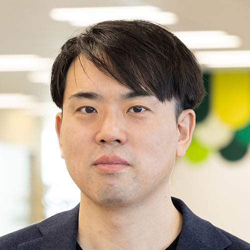 米田の顔のクローズアップ写真。短い黒髪を横分けに整え、落ち着いた表情で正面を見つめている。ネイビーのジャケットと黒のシャツを着用し、背景は柔らかくぼけた室内空間である