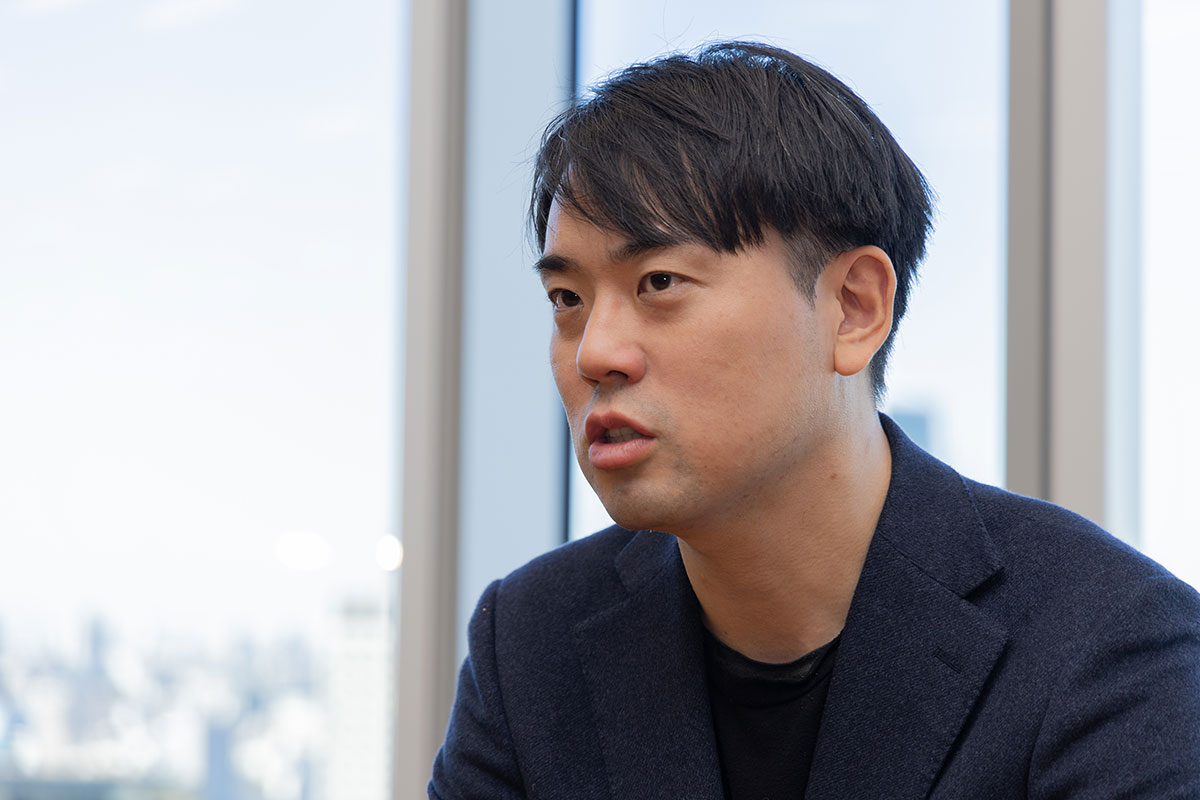 窓際の席で身を乗り出しながら話す米田の写真。ネイビーのジャケット姿で、真剣な表情を浮かべ横を向いている。背後には大きな窓があり、都市の景色が淡く広がっている