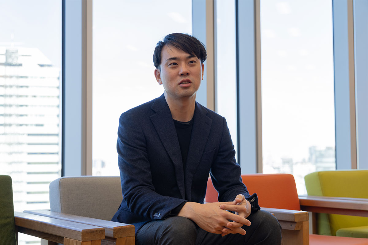 ソファに腰掛け、両手を組みながら話す米田の写真。ネイビージャケットと黒のインナーを着用し、落ち着いた表情で前方を見ている。背後には大きな窓と明るいオフィス空間が広がる