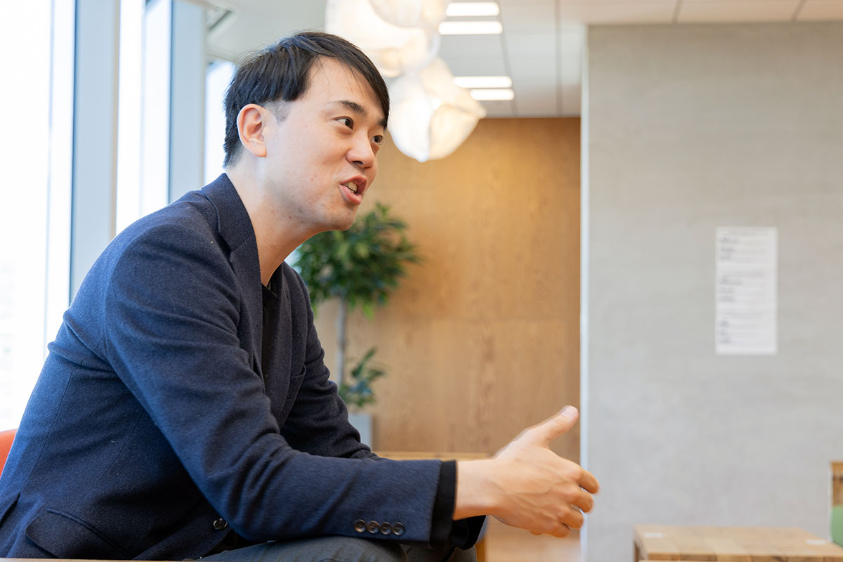 室内で身振りを交えながら話す米田の横顔写真。ネイビーのジャケットを着て前傾姿勢で手を差し出し、いきいきとした表情で会話している。背後には観葉植物と壁面が見える