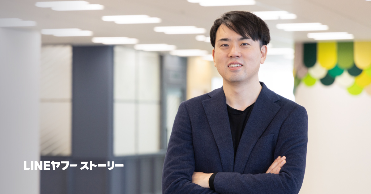 オフィス空間で腕を組み、正面を向いて立つ米田の写真。ネイビーのジャケットに黒のインナーを合わせ、短く整えた黒髪で穏やかに微笑んでいる。背後には明るい照明とカラフルな装飾が見える。