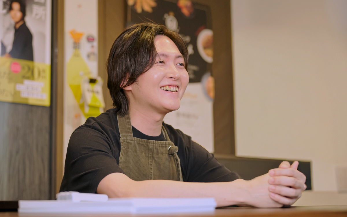 カウンターに両腕を置き、笑顔で話しているリュウジさんの様子。黒い服とエプロン姿で、リラックスした雰囲気がある。背景にはポスターが貼られ、和やかな対話の場面が写っている