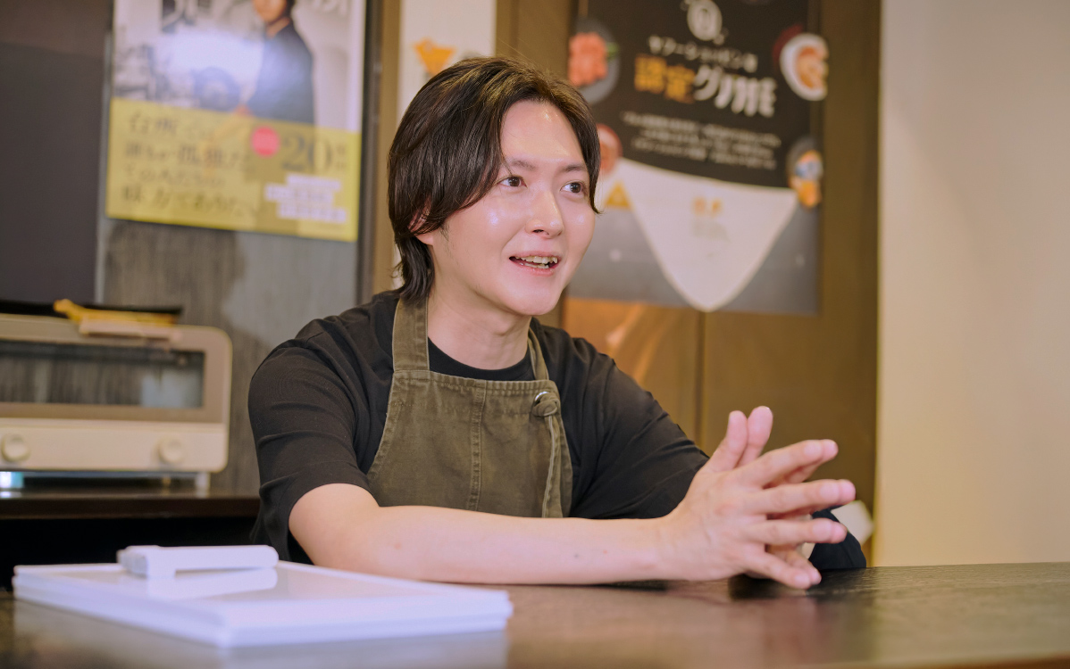店舗のカウンターに座り、身振りを交えながら話しているリュウジさんの様子。黒い服にカーキ色のエプロンを着用している。背景にはポスターや調理家電が並び、対談や取材の場面であることが伝わる