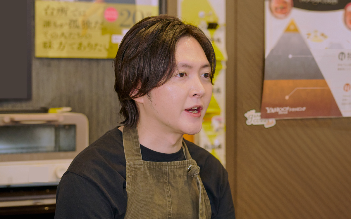 横向きの構図で、リュウジさんが話を聞くような表情をしている。黒髪を耳にかけ、カーキ色のエプロンを着ている。背景には掲示物や調理器具があり、室内でのインタビュー風景である