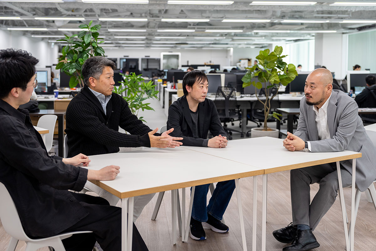 オフィスの一角で4人の男性がテーブルを囲んで会話している様子を写しています