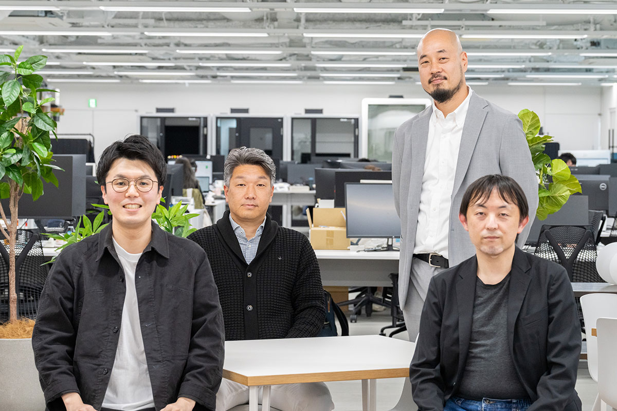 オフィスで4人の男性が集まり、カメラに向かってポーズをとっています。リラックスした雰囲気が伝わります