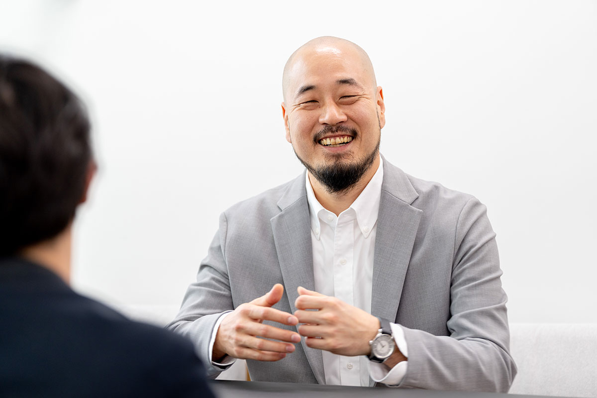 グレーのスーツを着た渡邊さんが笑顔で話しているシーンを捉えています