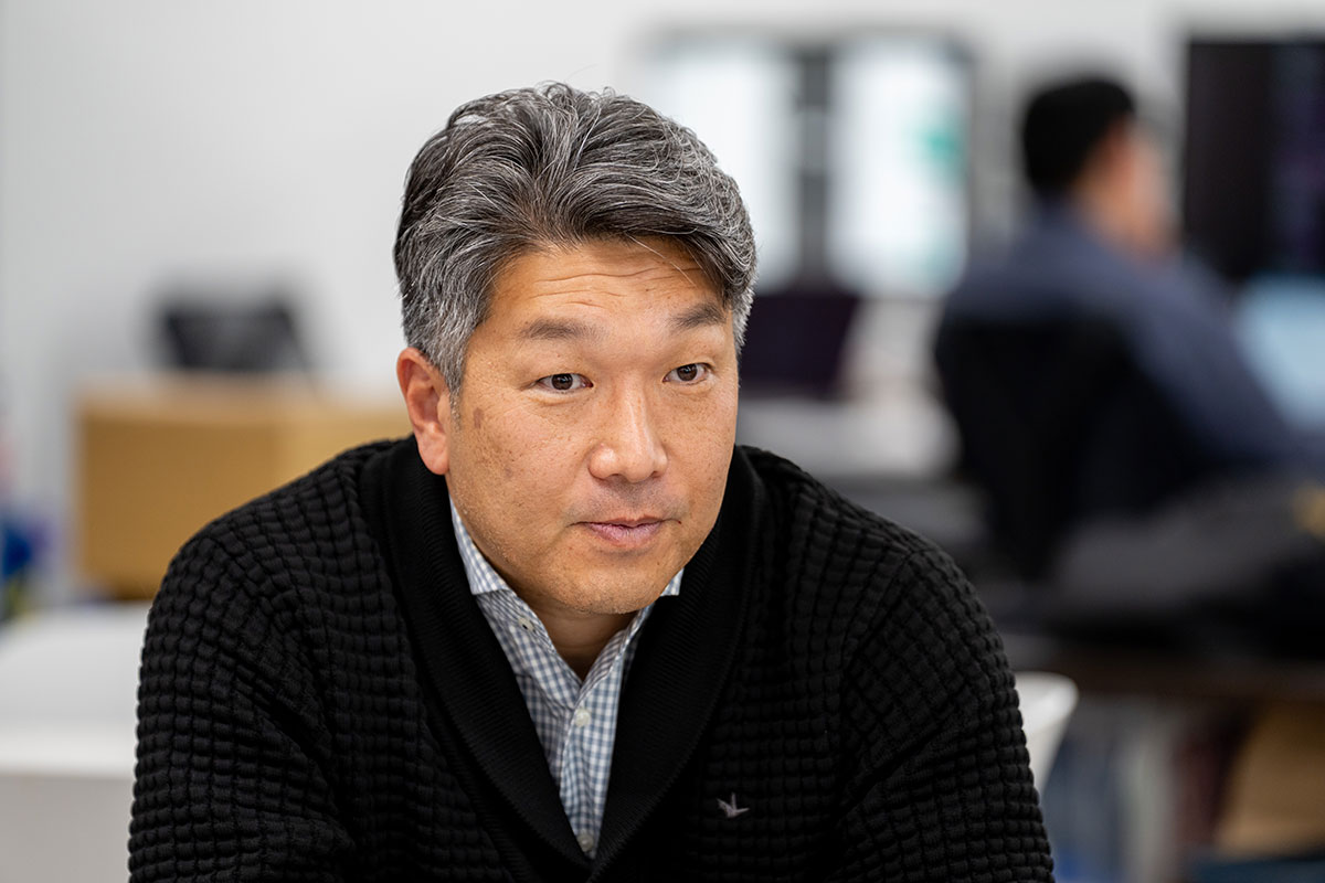 黒のカーディガンを着た福留が真剣な表情で前を見ている様子を捉えています。背景にはオフィスの一部が見えます