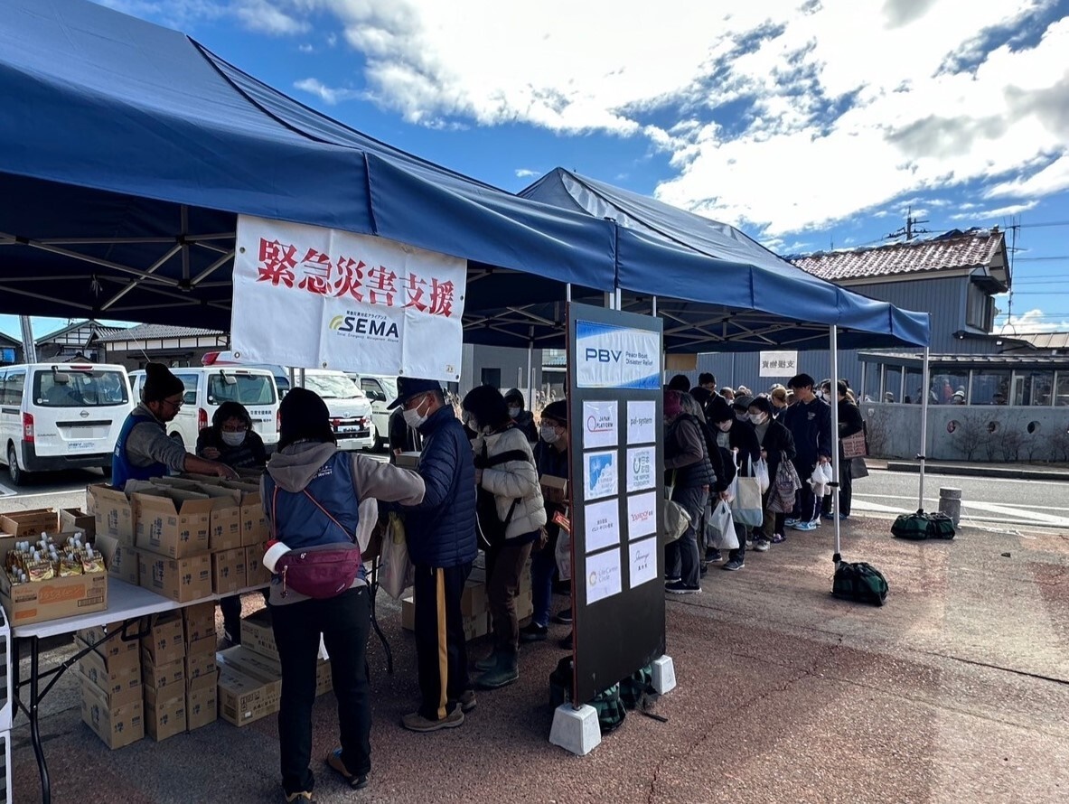 「緊急災害支援 緊急災害対応アライアンス SEMA」と掲げられた青いテントで、人々が箱詰めの救援物資を受け取っている様子を写した写真です。