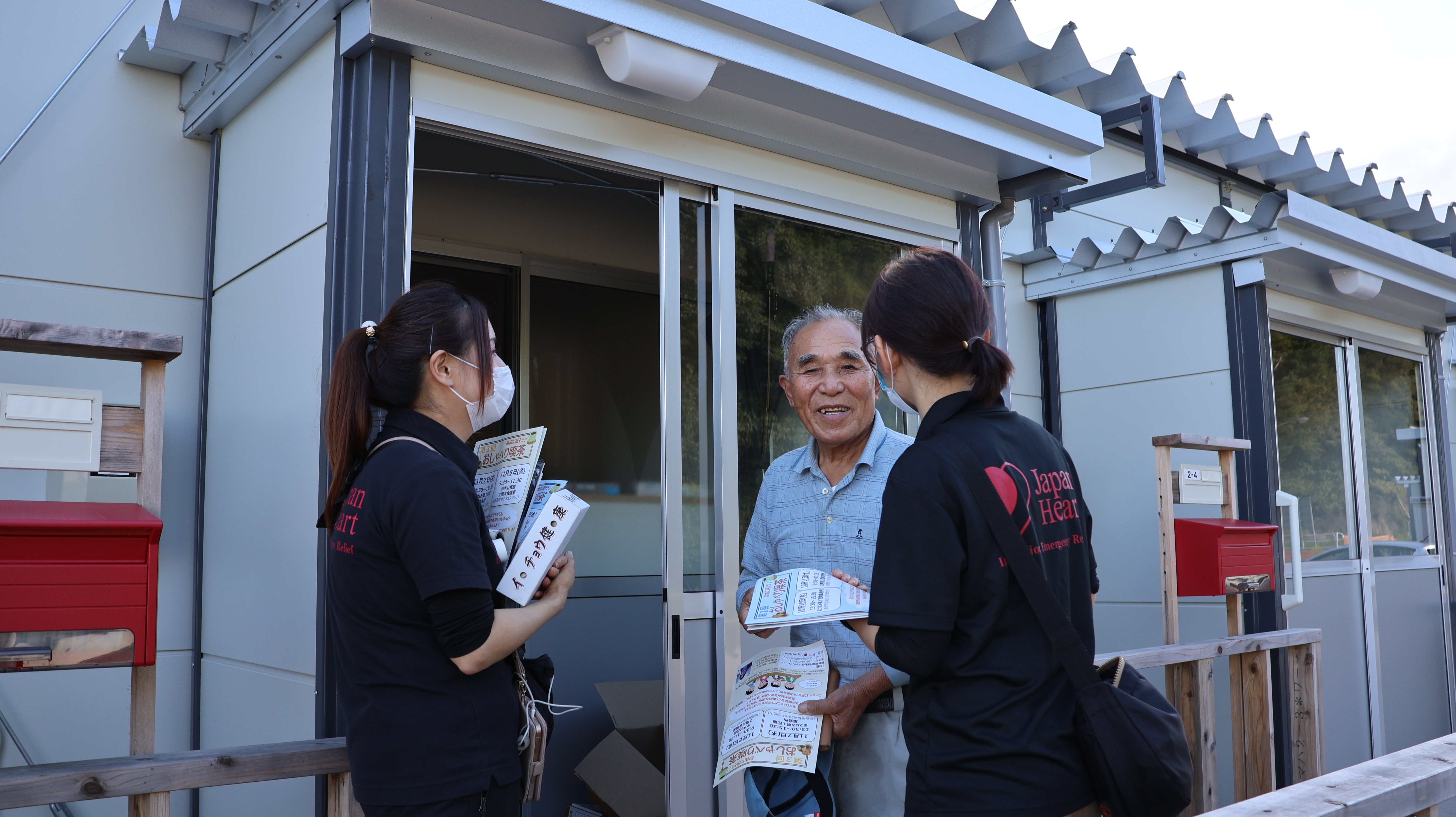 特定非営利活動法人ジャパンハートのメンバー2人が、仮設住宅に訪問し住人と会話するようすを写した写真です。