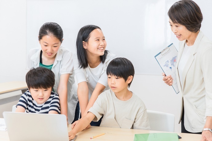 教室でノートパソコンを一緒に見る子どもたちと、クリップボードを持った女性が話しながら指導している様子です。
