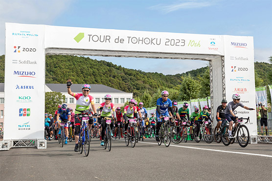 「TOUR de TOHOKU 2023 10th」と書かれたゲートをくぐり、多くのサイクリストがスタートを切る様子です。